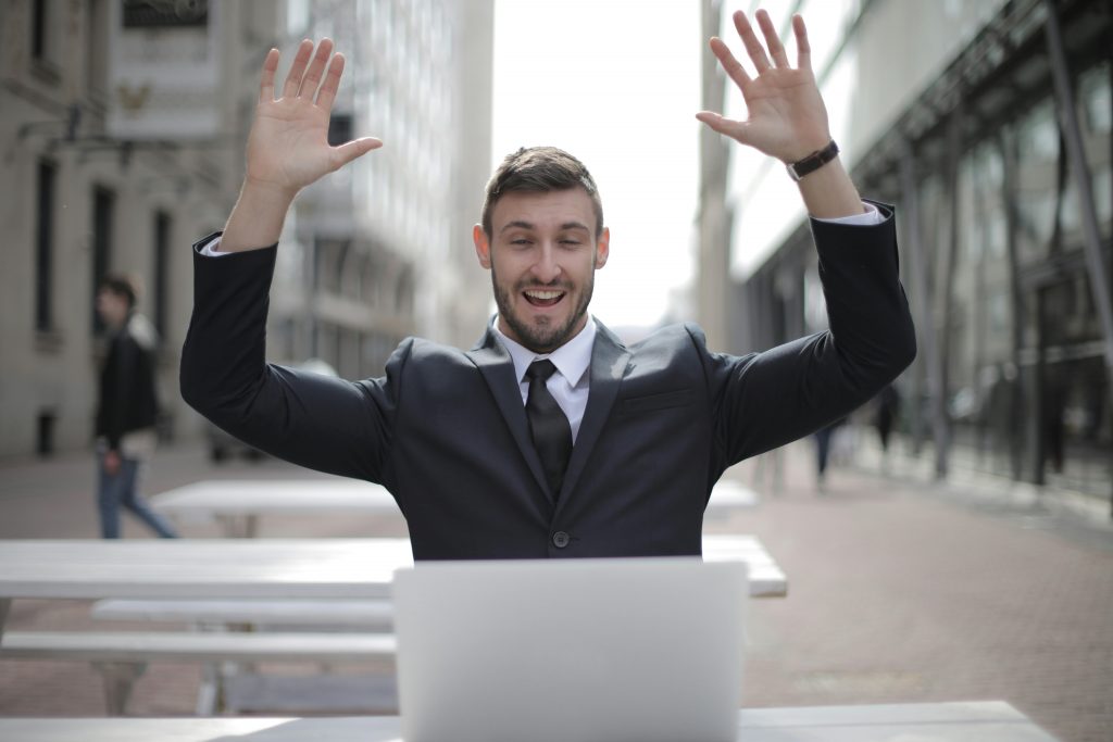 man in business attire smiling and raising his arms in celebration while looking at a laptop screen