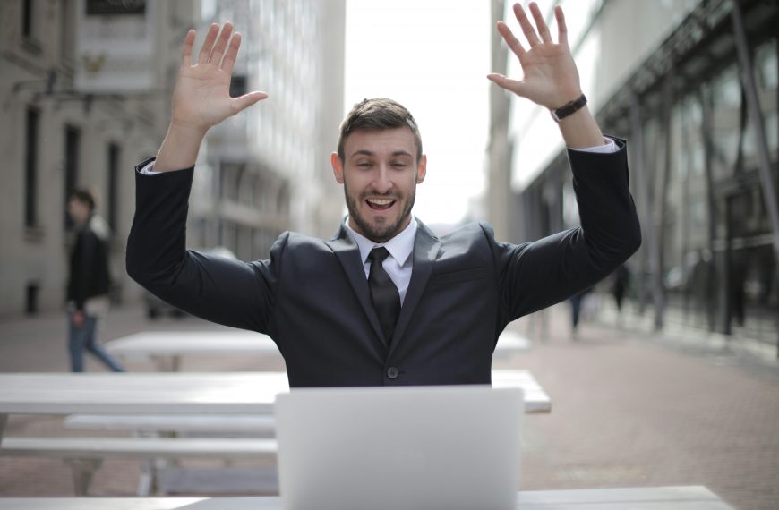 man in business attire smiling and raising his arms in celebration while looking at a laptop screen
